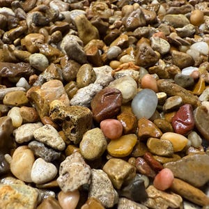 May include: A close-up view of a collection of smooth, polished river rocks. The stones vary in size, shape, and color, including shades of brown, tan, gray, and hints of red and pink. Some rocks have a glossy, wet appearance.