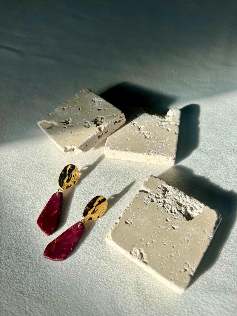 May include: Dangle earrings with gold-coloured, textured tops and burgundy-coloured, teardrop-shaped bottoms. The earrings are displayed with three off-white, square-shaped stone blocks on a white surface. The lighting creates shadows.