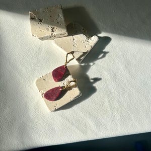 Peut inclure: Une paire de boucles d'oreilles dor&eacute;es avec des pendentifs en forme de goutte d'eau rouge fonc&eacute;. Les boucles d'oreilles sont pr&eacute;sent&eacute;es sur une surface claire et textur&eacute;e, avec quelques autres morceaux du m&ecirc;me mat&eacute;riau &eacute;parpill&eacute;s.
