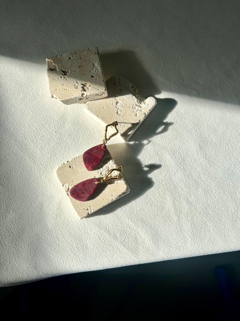 Peut inclure: Une paire de boucles d'oreilles dor&eacute;es avec des pendentifs en forme de goutte d'eau rouge fonc&eacute;. Les boucles d'oreilles sont pr&eacute;sent&eacute;es sur une surface claire et textur&eacute;e, avec quelques autres morceaux du m&ecirc;me mat&eacute;riau &eacute;parpill&eacute;s.