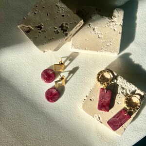 May include: Two pairs of earrings displayed on textured, off-white stone blocks. One pair features gold square tops with round, red marbled drops. The other pair has gold hoops with rectangular, red marbled pendants. The earrings are set against a neutral background.