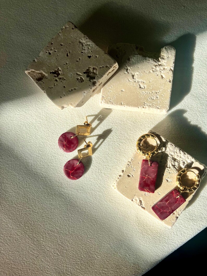 May include: Two pairs of earrings displayed on textured, off-white stone blocks. One pair features gold square tops with round, red marbled drops. The other pair has gold hoops with rectangular, red marbled pendants. The earrings are set against a neutral background.