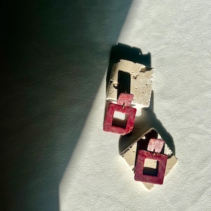 May include: Two pairs of earrings. Each earring features a square, open-center design in a deep red hue with a marbled effect. The earrings are attached to a smaller, rectangular piece of off-white material.