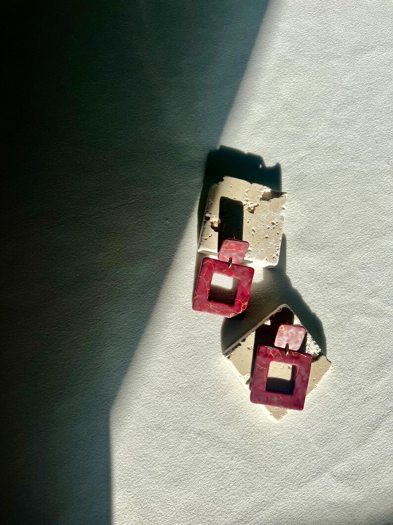 May include: Two pairs of earrings. Each earring features a square, open-center design in a deep red hue with a marbled effect. The earrings are attached to a smaller, rectangular piece of off-white material.