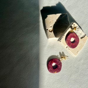 May include: A pair of burgundy and gold earrings with a square and teardrop shape. The earrings are displayed on a small, textured, off-white surface. The earrings have gold-colored posts.