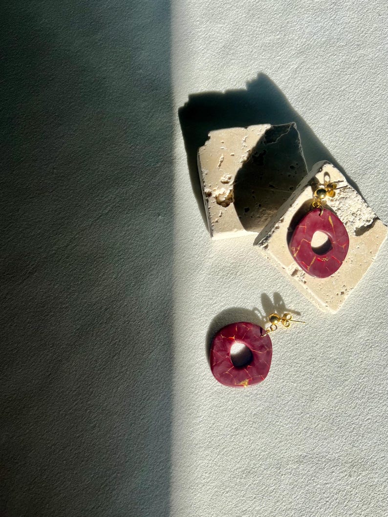 May include: A pair of burgundy and gold earrings with a square and teardrop shape. The earrings are displayed on a small, textured, off-white surface. The earrings have gold-colored posts.