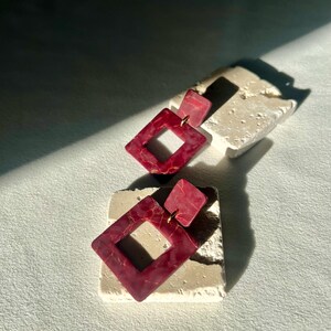 May include: Burgundy geometric earrings with a gold marbled effect. Each earring has a small square stud and a larger diamond-shaped pendant. The earrings are displayed on a textured, off-white surface.