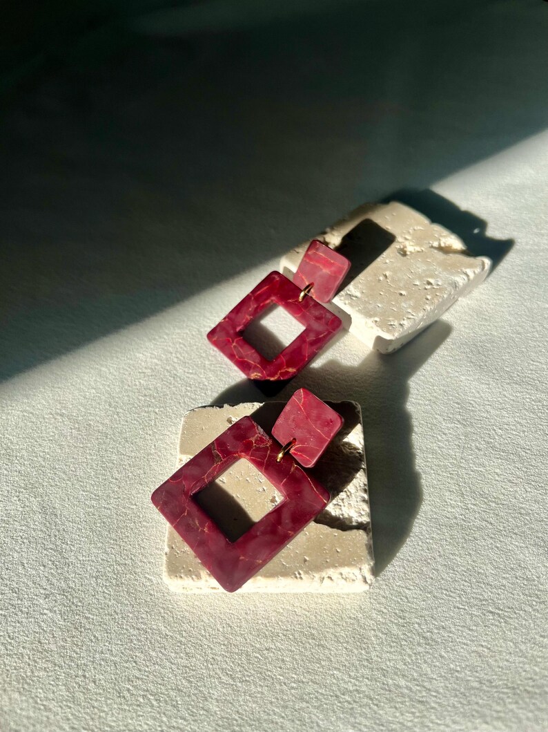 May include: A pair of burgundy geometric earrings with a marbled gold pattern. Each earring features a small square stud and a larger diamond-shaped pendant. The earrings are displayed on a textured, off-white surface.