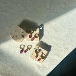 May include: Three pairs of gold earrings with pink and purple detailing are shown on textured, off-white blocks. The earrings feature square hoops, teardrop dangles, and circular hoops. They are displayed on a white surface, with shadows cast across the scene.