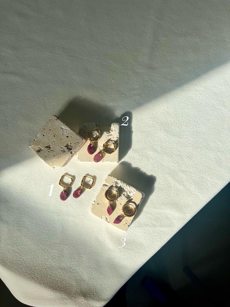 May include: Three pairs of gold earrings with pink and purple detailing are shown on textured, off-white blocks. The earrings feature square hoops, teardrop dangles, and circular hoops. They are displayed on a white surface, with shadows cast across the scene.