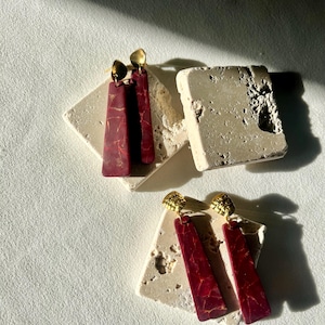 K&ouml;nnte beinhalten: Zwei Paar Ohrh&auml;nger. Jeder Ohrring hat einen rechteckigen, weinroten Anh&auml;nger mit goldenen Marmorierungen, der von einem kleinen, strukturierten goldenen Ohrstecker gekr&ouml;nt wird. Die Ohrringe liegen auf hellfarbigen, strukturierten Steinplattformen.