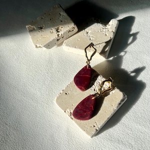 May include: Dangle earrings featuring a deep red, teardrop-shaped design, suspended from gold-toned geometric frames. The earrings are displayed on a textured, off-white surface, with shadows from natural light.
