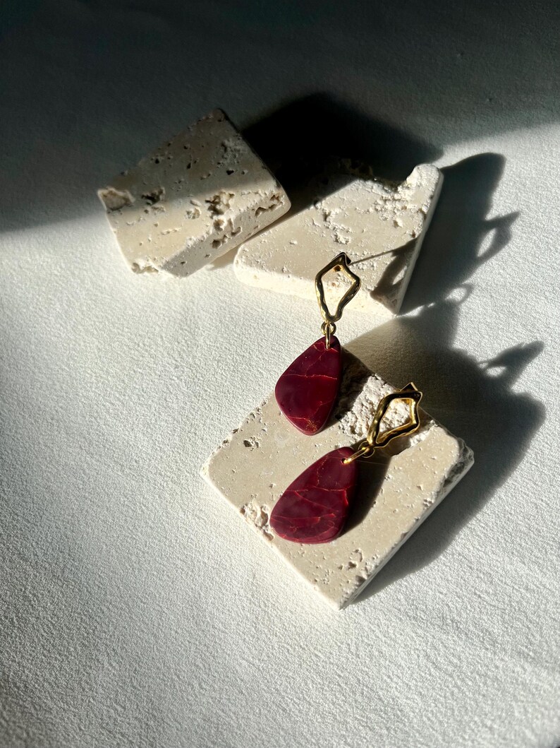 Peut inclure: Boucles d'oreilles pendantes avec une forme de goutte d'eau rouge fonc&eacute;, fix&eacute;es sur des montures g&eacute;om&eacute;triques dor&eacute;es. Elles reposent sur une surface textur&eacute;e blanc cass&eacute;, avec des ombres naturelles.