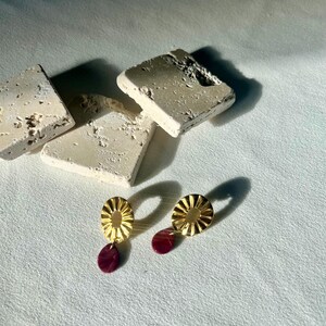 May include: Gold-coloured earrings featuring a sunburst design and a burgundy teardrop-shaped dangle. The earrings are presented on a white surface, with textured, off-white stone blocks in the background.