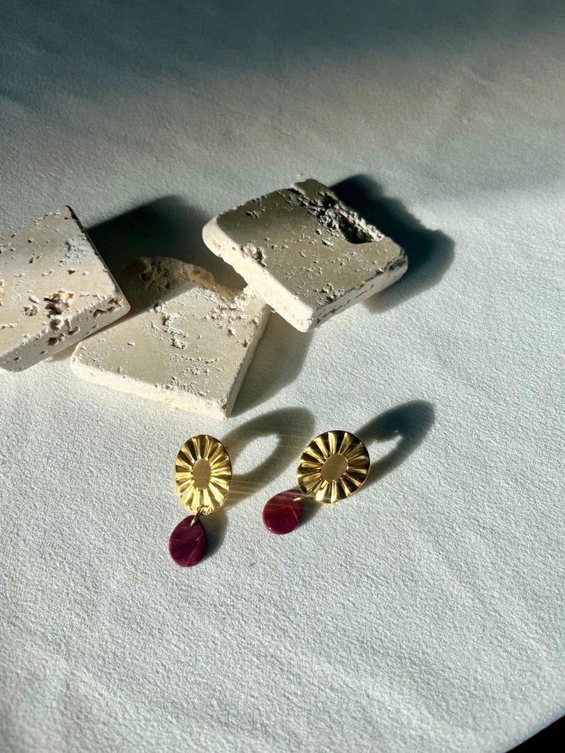 Pu&ograve; includere: Un paio di orecchini color oro con un design a forma di sole e un pendente a goccia color bordeaux. Gli orecchini sono esposti su una superficie bianca con blocchi di pietra strutturati bianco sporco sullo sfondo.