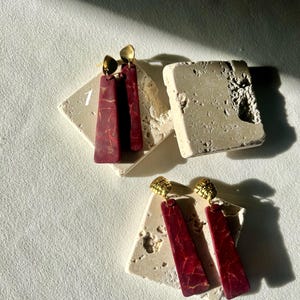 K&ouml;nnte beinhalten: Zwei Paar Ohrringe. Ein Paar hat einen rechteckigen, weinroten Anh&auml;nger mit goldenen Akzenten und einen goldfarbenen Stecker. Das zweite Paar hat ein &auml;hnliches Design. Beide Paare werden auf einer strukturierten, cremefarbenen Oberfl&auml;che pr&auml;sentiert.