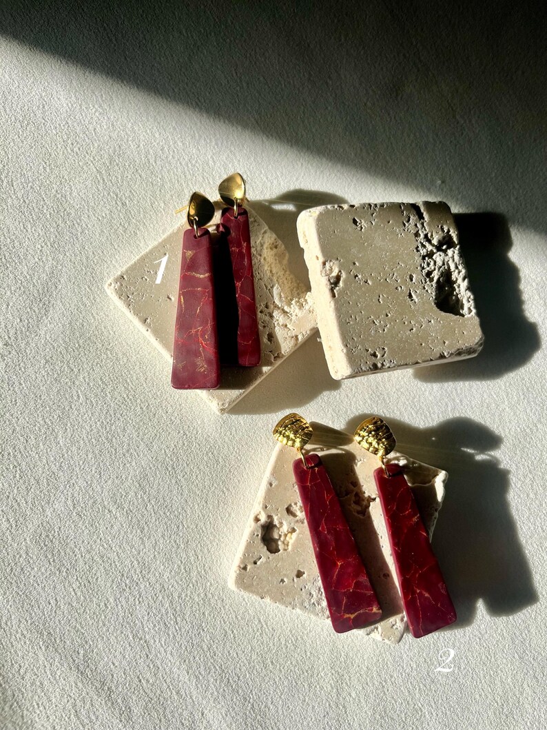 K&ouml;nnte beinhalten: Zwei Paar Ohrringe. Ein Paar hat einen rechteckigen, weinroten Anh&auml;nger mit goldenen Akzenten und einen goldfarbenen Stecker. Das zweite Paar hat ein &auml;hnliches Design. Beide Paare werden auf einer strukturierten, cremefarbenen Oberfl&auml;che pr&auml;sentiert.