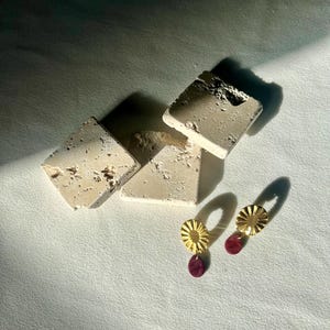 Pu&ograve; includere: Orecchini dorati a forma di sole con un piccolo pendente rotondo in pietra rosso scuro. Gli orecchini sono esposti su una superficie bianca accanto a diversi blocchi rettangolari beige strutturati.