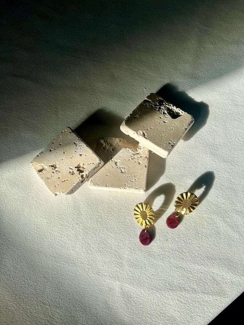 Pu&ograve; includere: Orecchini dorati a forma di sole con un piccolo pendente rotondo in pietra rosso scuro. Gli orecchini sono esposti su una superficie bianca accanto a diversi blocchi rettangolari beige strutturati.