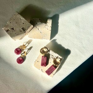May include: A pair of gold-colored hoop earrings with rectangular, burgundy-colored pendants and a pair of gold-colored square stud earrings with round, burgundy-colored pendants. The earrings are displayed on a textured, off-white surface.
