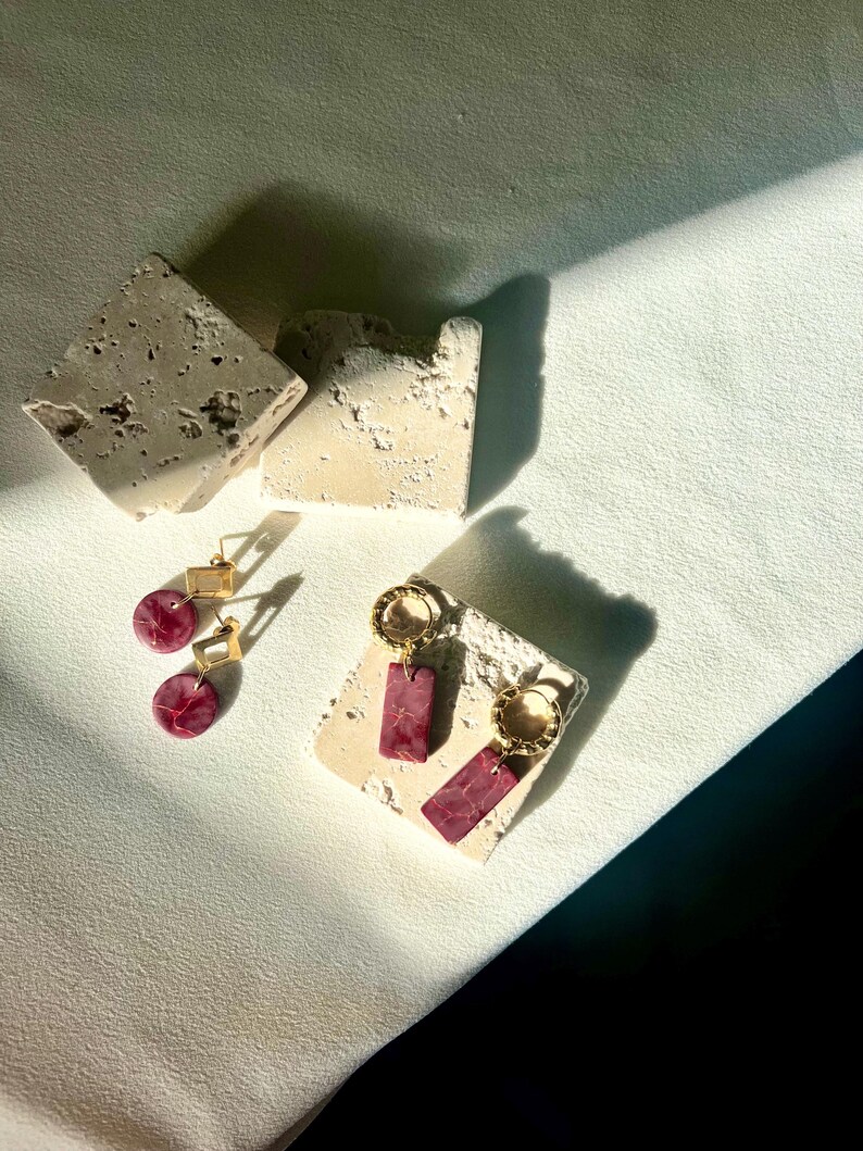 May include: A pair of gold-colored hoop earrings with rectangular, burgundy-colored pendants and a pair of gold-colored square stud earrings with round, burgundy-colored pendants. The earrings are displayed on a textured, off-white surface.
