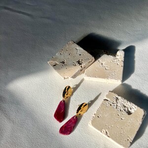 May include: Earrings with gold tops and burgundy pendants, displayed on a white surface with textured stone blocks. The pendants have a teardrop shape. The stone blocks are off-white in colour.