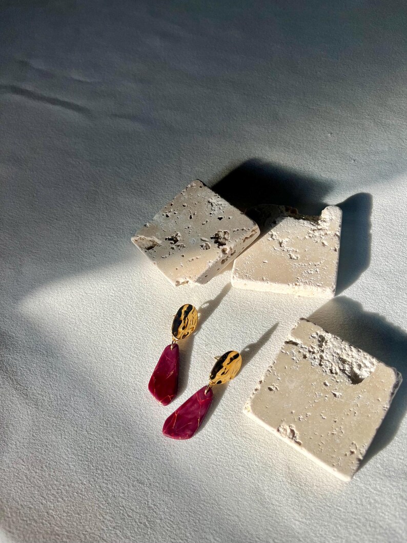 May include: Earrings with gold tops and burgundy pendants, displayed on a white surface with textured stone blocks. The pendants have a teardrop shape. The stone blocks are off-white in colour.