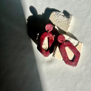 May include: A pair of red and gold geometric earrings. The earrings have a marbled effect and a unique shape with a circular top and an open, angular bottom. The earrings are displayed on a textured, off-white surface.
