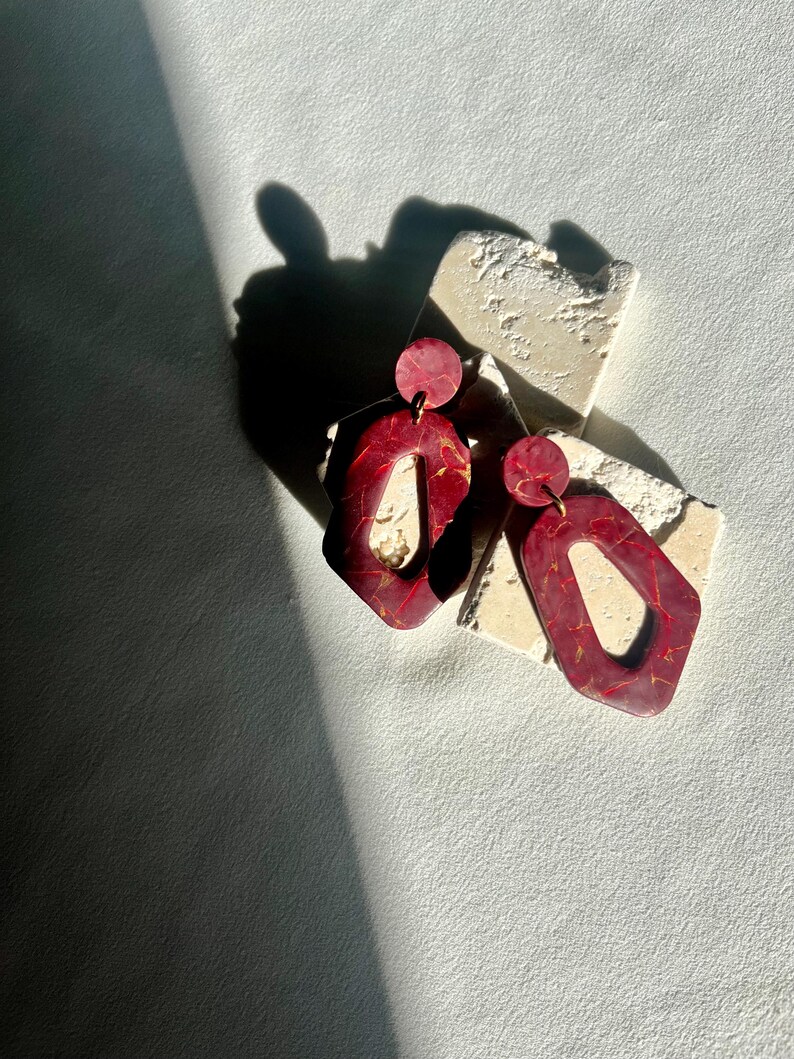 May include: A pair of red and gold geometric earrings. The earrings have a marbled effect and a unique shape with a circular top and an open, angular bottom. The earrings are displayed on a textured, off-white surface.