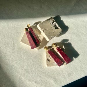 K&ouml;nnte beinhalten: Zwei Paar Ohrringe mit geometrischem Design. Die Ohrringe haben einen goldfarbenen Stecker und einen langen, rechteckigen Anh&auml;nger in tiefroter Farbe mit Marmormuster. Die Ohrringe werden auf kleinen, quadratischen, cremefarbenen Steinbl&ouml;cken pr&auml;sentiert.