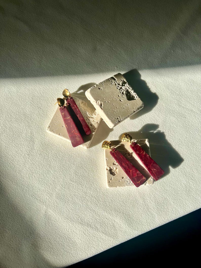 K&ouml;nnte beinhalten: Zwei Paar Ohrringe mit geometrischem Design. Die Ohrringe haben einen goldfarbenen Stecker und einen langen, rechteckigen Anh&auml;nger in tiefroter Farbe mit Marmormuster. Die Ohrringe werden auf kleinen, quadratischen, cremefarbenen Steinbl&ouml;cken pr&auml;sentiert.