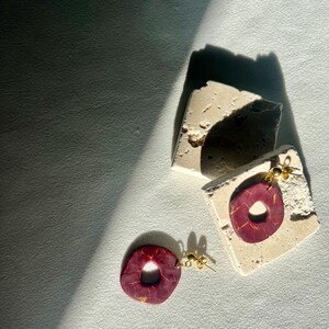 Puede incluir: Pendientes hechos a mano con forma de rosquilla, en color granate con vetas doradas. Cuelgan de postes dorados y se exhiben sobre una superficie texturizada de color blanquecino.