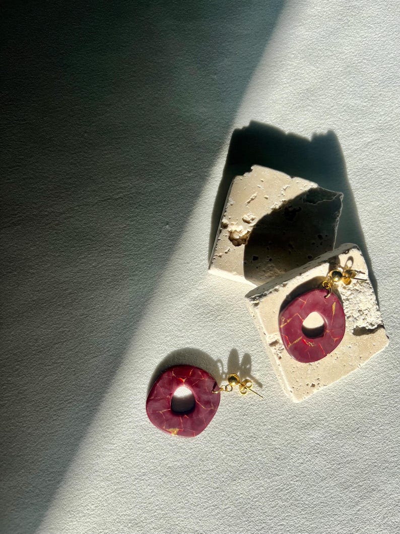May include: A pair of handmade earrings with a maroon and gold marbled design. The earrings are donut-shaped and hang from gold-colored posts. They are displayed on a textured, off-white surface.