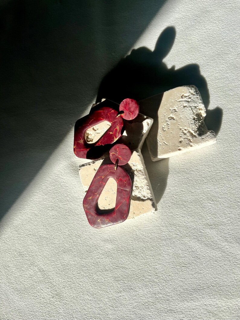 May include: Burgundy and gold-veined resin earrings. The earrings have a geometric design with a circular stud and a larger, open-shape pendant. The earrings are displayed on a textured, off-white surface.