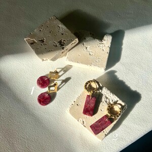 May include: Two pairs of gold-toned earrings with red marbled accents. One pair features a square top and a circular bottom, while the other has a circular hoop and rectangular drops. The earrings are displayed on textured, off-white blocks.