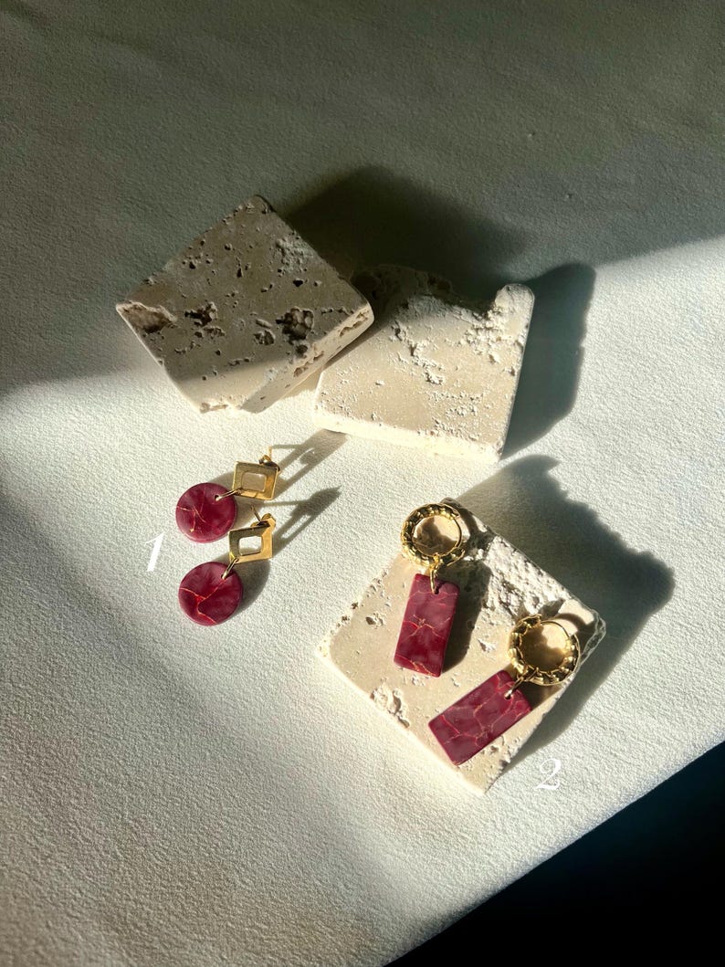 May include: Two pairs of gold-toned earrings with red marbled accents. One pair features a square top and a circular bottom, while the other has a circular hoop and rectangular drops. The earrings are displayed on textured, off-white blocks.