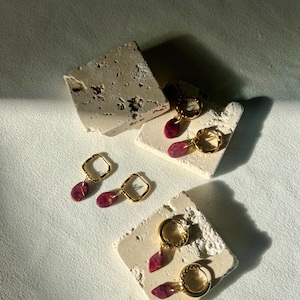 Puede incluir: Pendientes de aro dorados con un colgante de piedra en forma de lágrima de color burdeos. Los pendientes se exhiben sobre pequeños bloques de piedra texturizada de color blanquecino. Diseño moderno y minimalista.