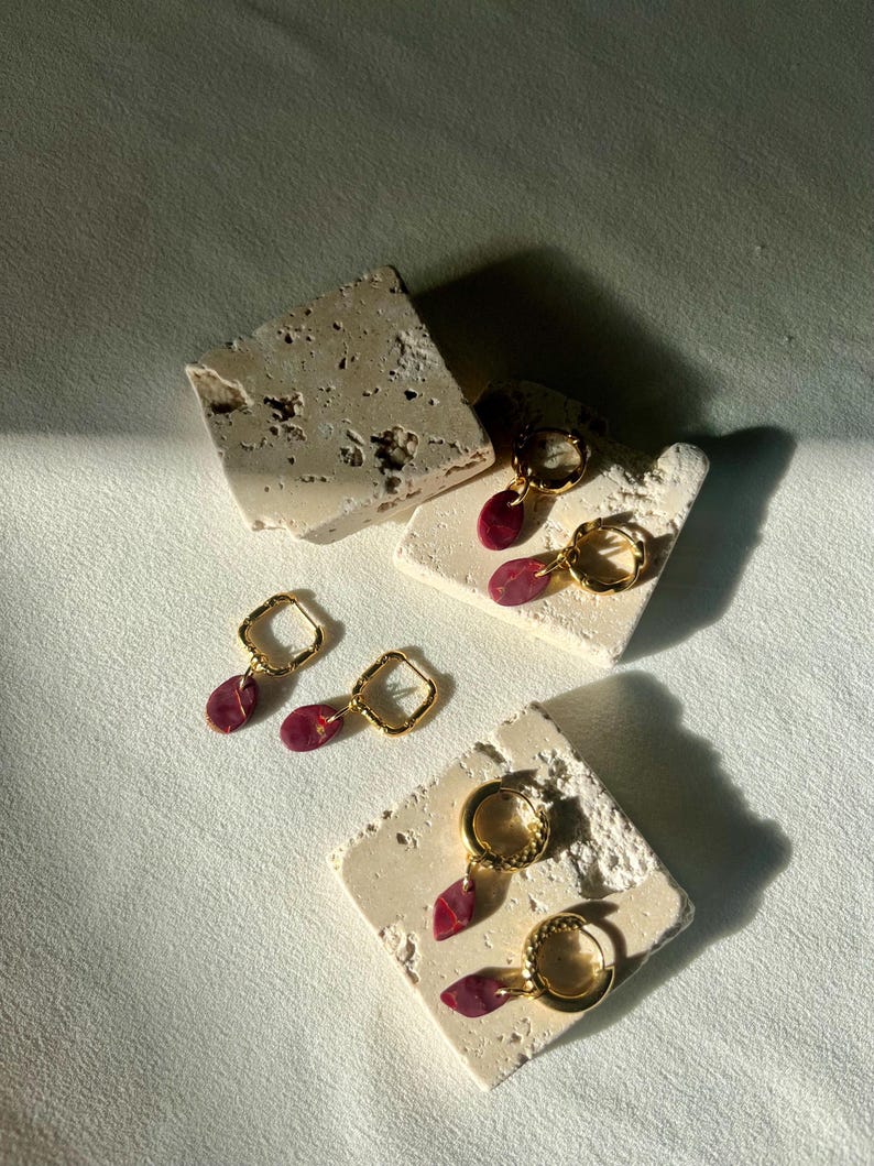 May include: Gold-coloured hoop earrings with a teardrop-shaped, burgundy-coloured stone pendant. The earrings are displayed on small, textured, off-white stone blocks. The earrings have a modern, minimalist design, and are suitable for everyday wear.