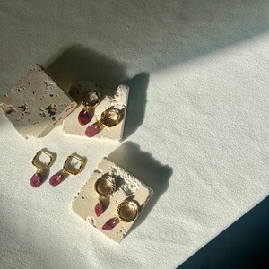 May include: Gold-coloured hoop and square earrings with teardrop-shaped, pink gemstones. The earrings are displayed on small, textured, off-white blocks, with a soft, white background and shadows.