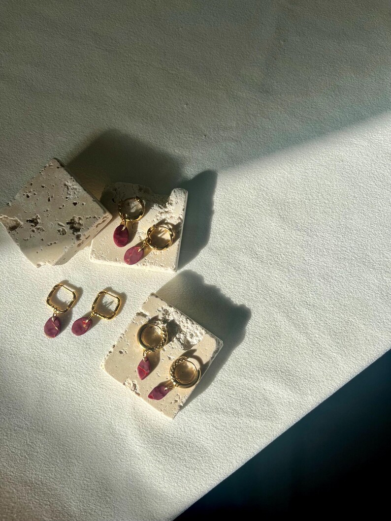 May include: Gold-coloured hoop and square earrings with teardrop-shaped, pink gemstones. The earrings are displayed on small, textured, off-white blocks, with a soft, white background and shadows.