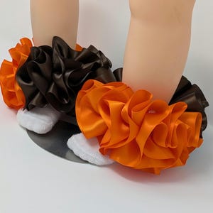 May include: A pair of baby leg warmers with ruffled orange and brown satin fabric. The leg warmers are worn over white socks. The background is a light color.