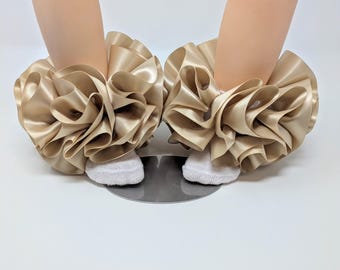 May include: Two pairs of gold satin ruffled ankle cuffs, each worn over a white sock. The cuffs are made of layered, flowing fabric, creating a voluminous, decorative effect. The background is a light, neutral color.