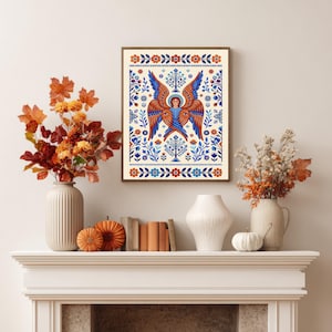 Może przedstawiać: Oprawiony wydruk przedstawiający anioła w stylu ludowym z niebieskimi i brązowymi skrzydłami, otoczony kwiatowymi wzorami w kolorze niebieskim, pomarańczowym i białym. Grafika jest eksponowana nad kominkiem.