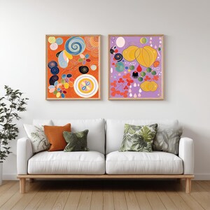 Peut inclure: Deux impressions d'art abstrait encadrées. L'une présente un motif tourbillonnant orange, bleu et jaune. L'autre présente des formes jaunes et oranges sur fond violet. Les impressions sont au-dessus d'un canapé blanc avec des coussins décoratifs.