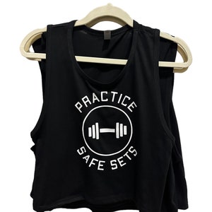 May include: Black crop top with the text "PRACTICE SAFE SETS" in white, featuring a barbell graphic inside a circle. The top is sleeveless and hangs on a beige hanger, suitable for gym workouts.