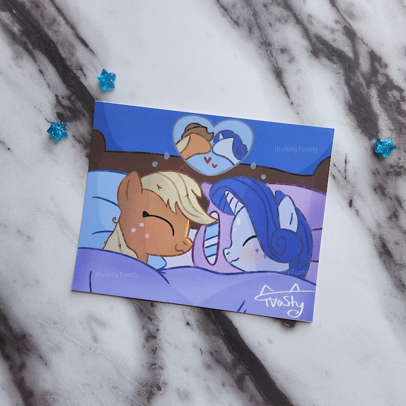 May include: A digital illustration of two cartoon ponies sleeping in a bed together. The ponies are a light brown pony with a blonde mane and a blue pony with a purple mane. The ponies are both smiling and have their eyes closed. The background is a blue and purple color scheme. The text "itty bitty toasty" is in the top right corner of the image.