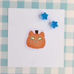May include: A white card with a cartoon orange pumpkin with a cat face on it. There are two blue glitter stars on the card.