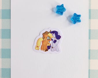 May include: A sticker featuring a cartoon image of two ponies, one with an orange and yellow coat and a cowboy hat, and the other with a purple and white coat and a purple mane. The ponies are facing each other and appear to be hugging.