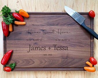 May include: A personalized walnut cutting board with the text "Home Sweet Home", coordinates, "James + Tessa", and "EST. 2022". The board is surrounded by vegetables and a knife.