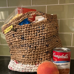 May include: A brown woven bag with a white and red trim, filled with groceries. A can of Campbell's Condensed Soup, Mushroom flavor, and a peach are sitting on a dark surface next to the bag.